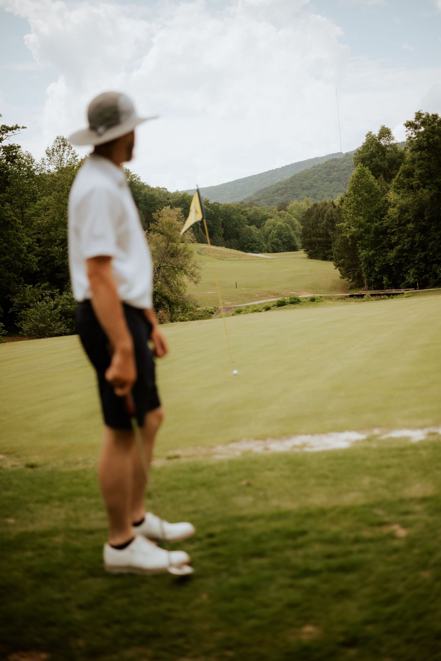 Golfer in white shirt and hat stands on lush green golf course with yellow flag and mountain backdrop.