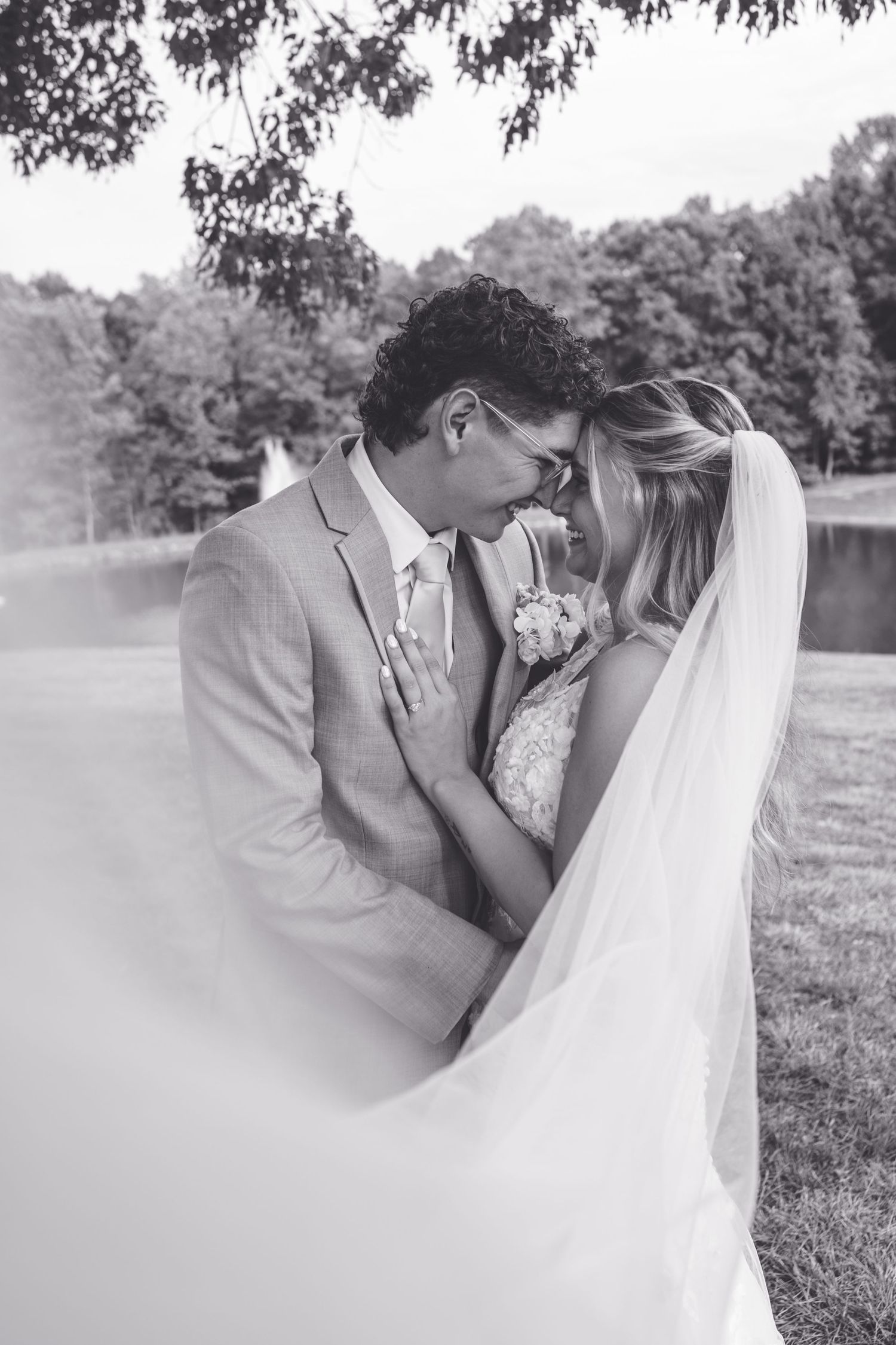 Black and white wedding photo of bride and groom sharing an intimate moment outdoors under trees.