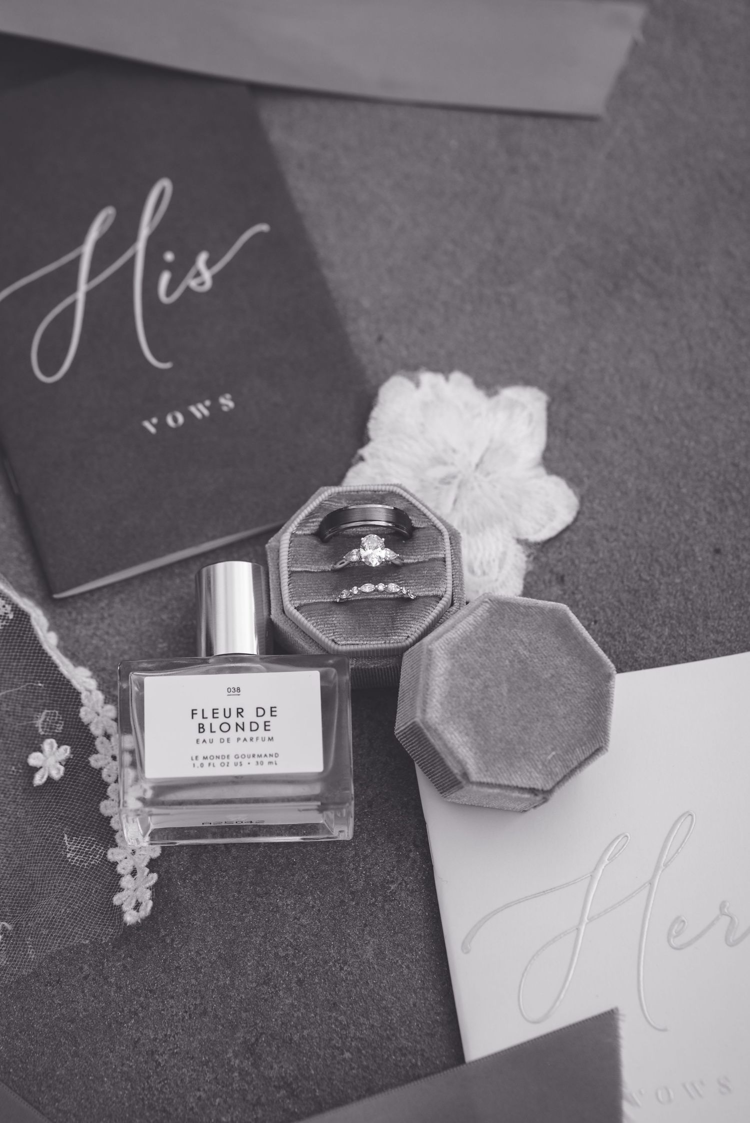 Black and white flat lay of wedding rings in velvet box, Fleur de Blonde perfume, and vow booklets.