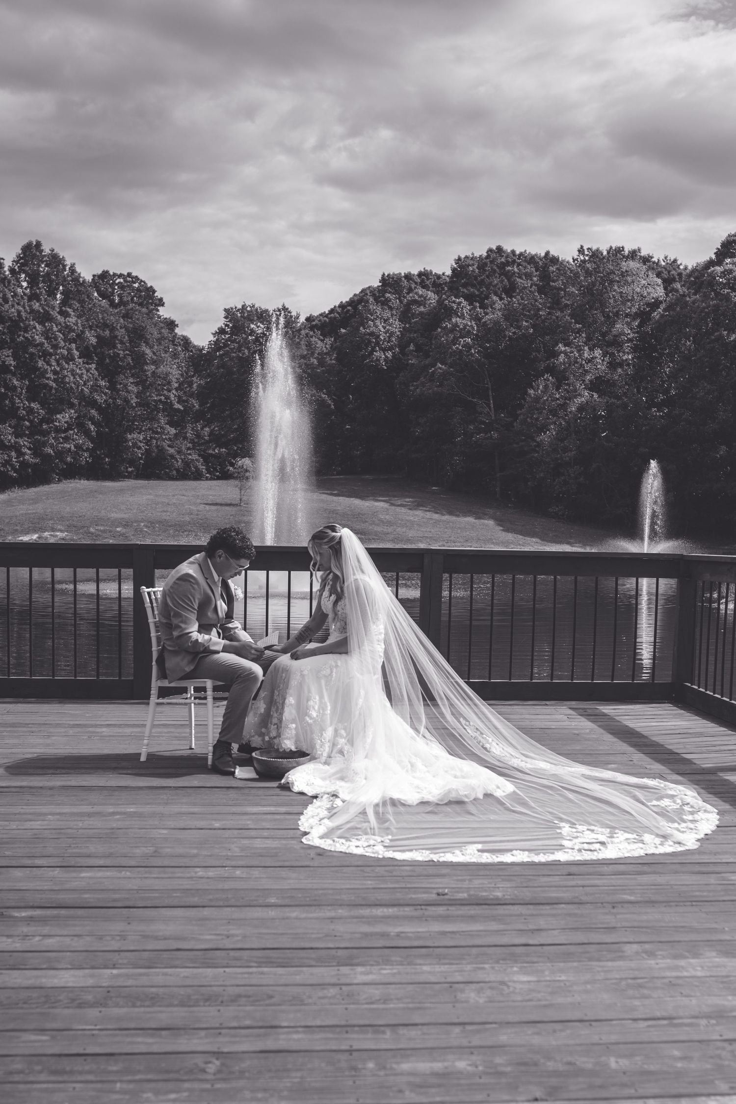 Black and white photo of bride and groom sitting on deck by fountain lake, bride's long veil trailing behind her.