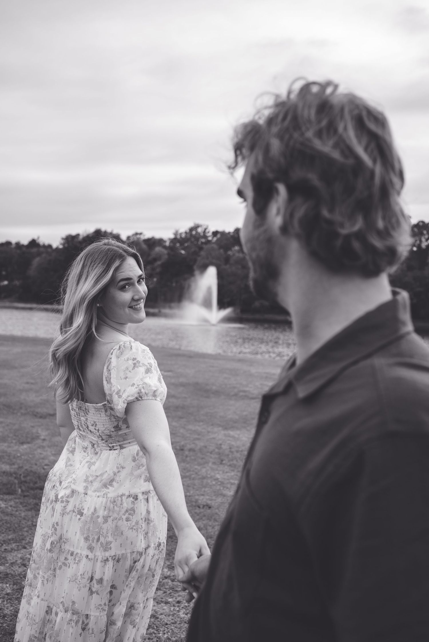Black and white photo of a smiling woman in a floral dress holding hands with a curly-haired man near a fountain.