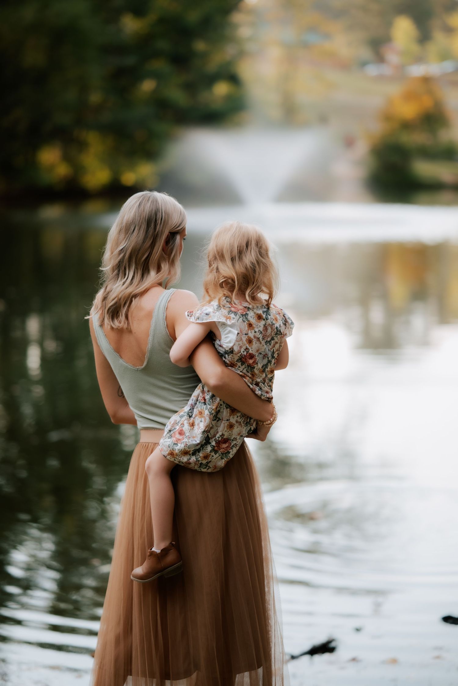 Figure holds small child while standing near pond with fountain in background.