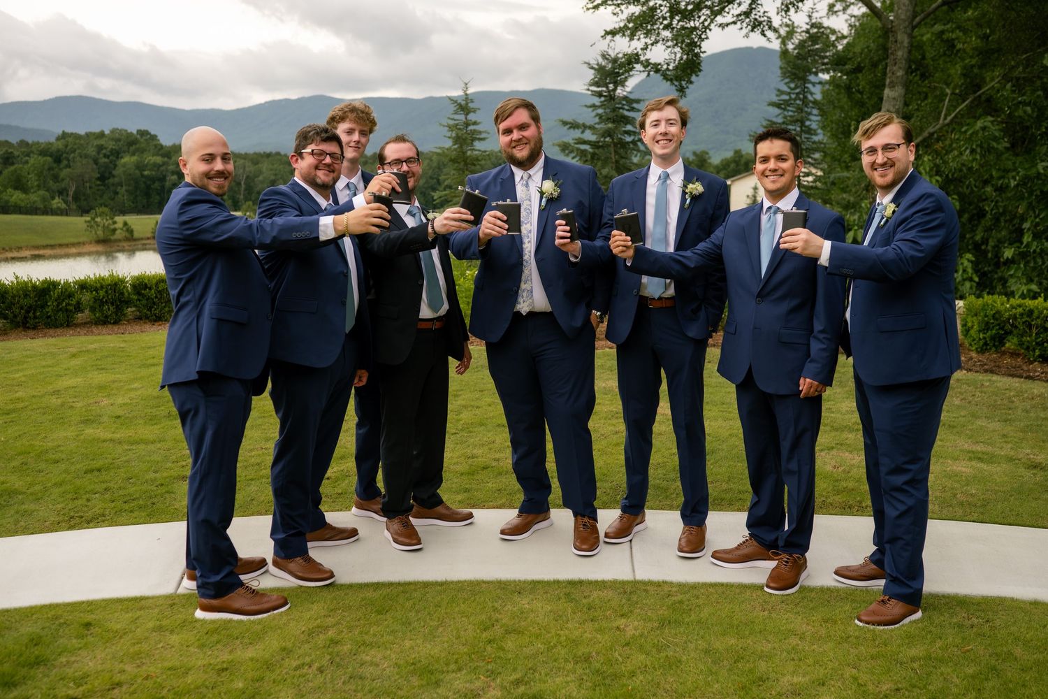 Eight groomsmen in navy suits toast with drinks outdoors at a wedding venue with mountain views.