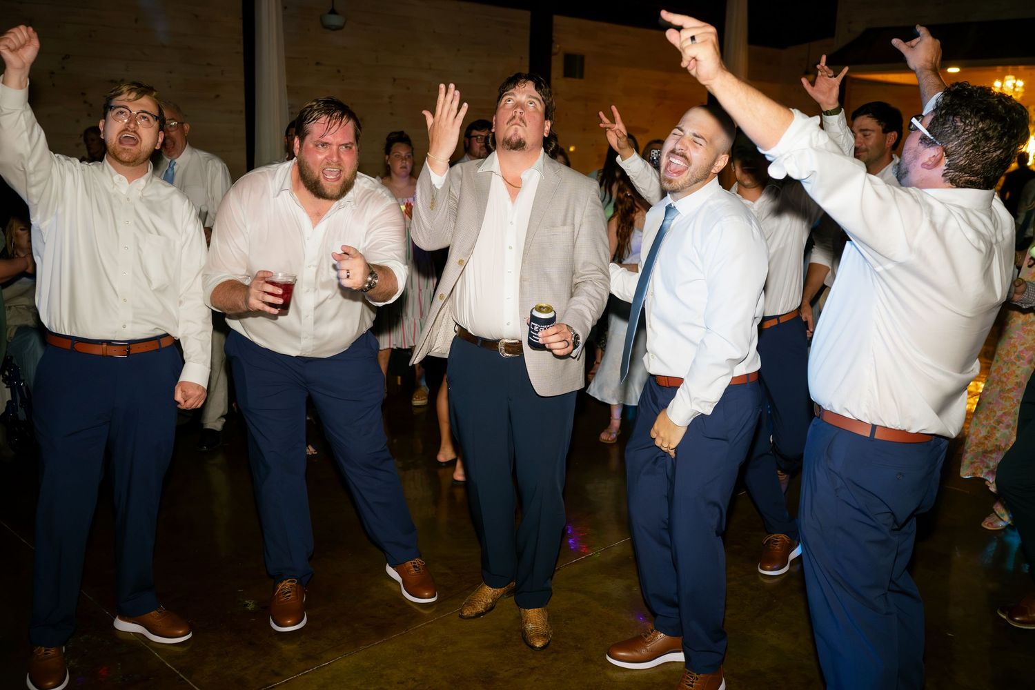 Group of groomsmen in navy pants and white shirts dancing joyfully at wedding reception.