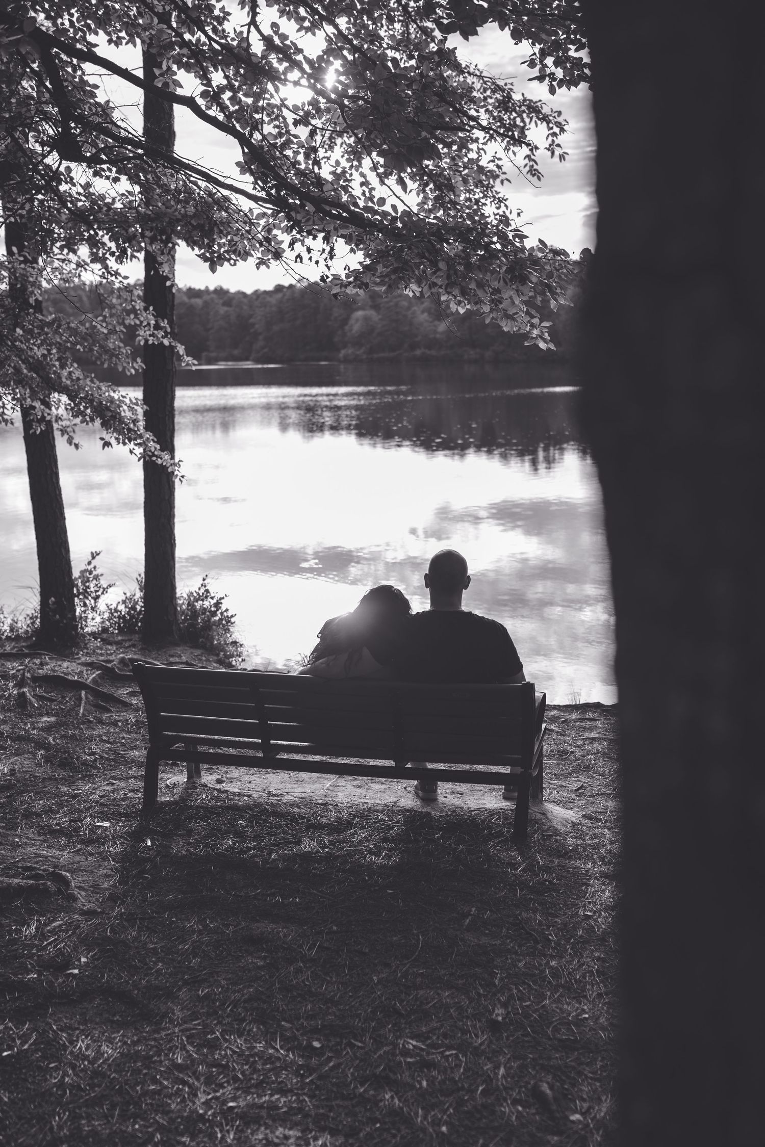 Black and white photo of a couple sitting on a bench by a tranquil lake, surrounded by trees.
