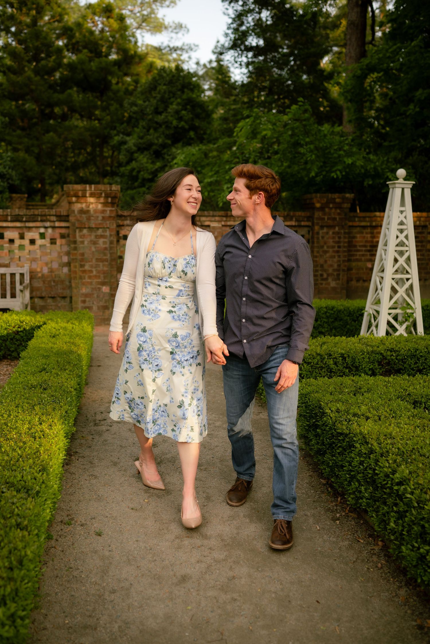 Couple walking hand in hand through a lush garden pathway, smiling at each other, woman in floral dress, man in dark shirt.