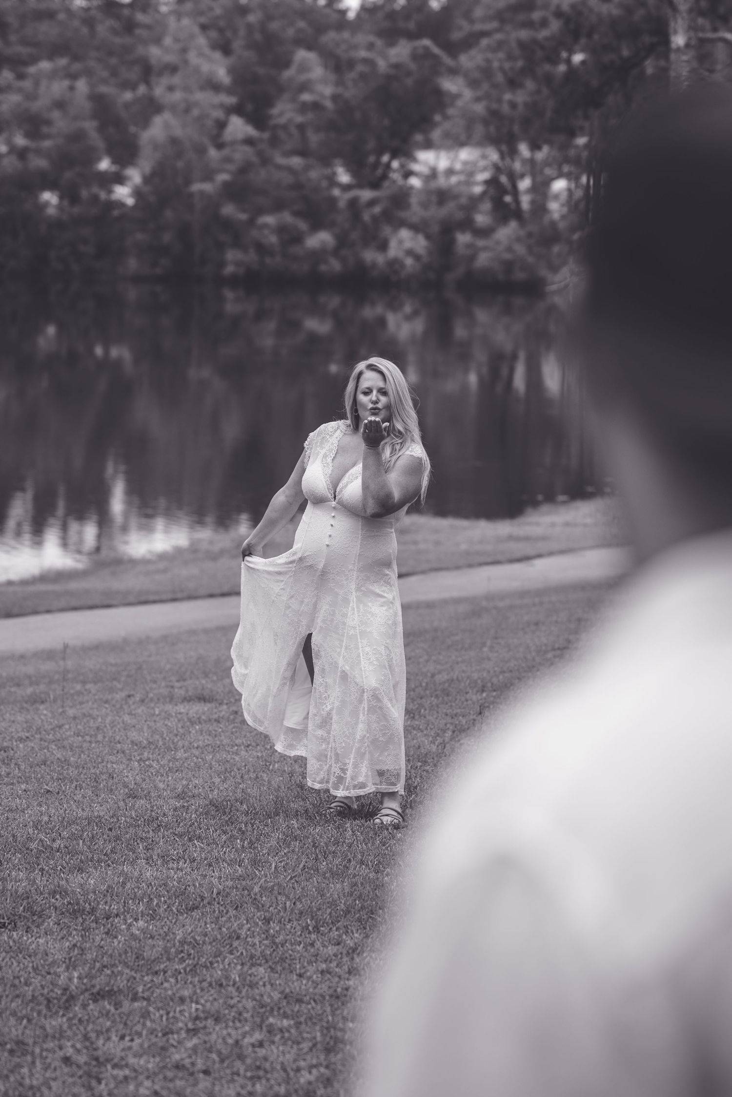 Black and white photo of a woman in a flowing white dress standing by a lake in a lush green park.