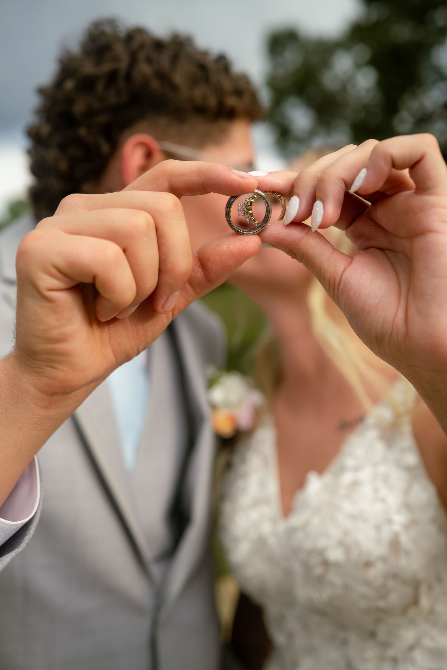 Bride and groom hold up matching gold wedding rings together outdoors on their wedding day.