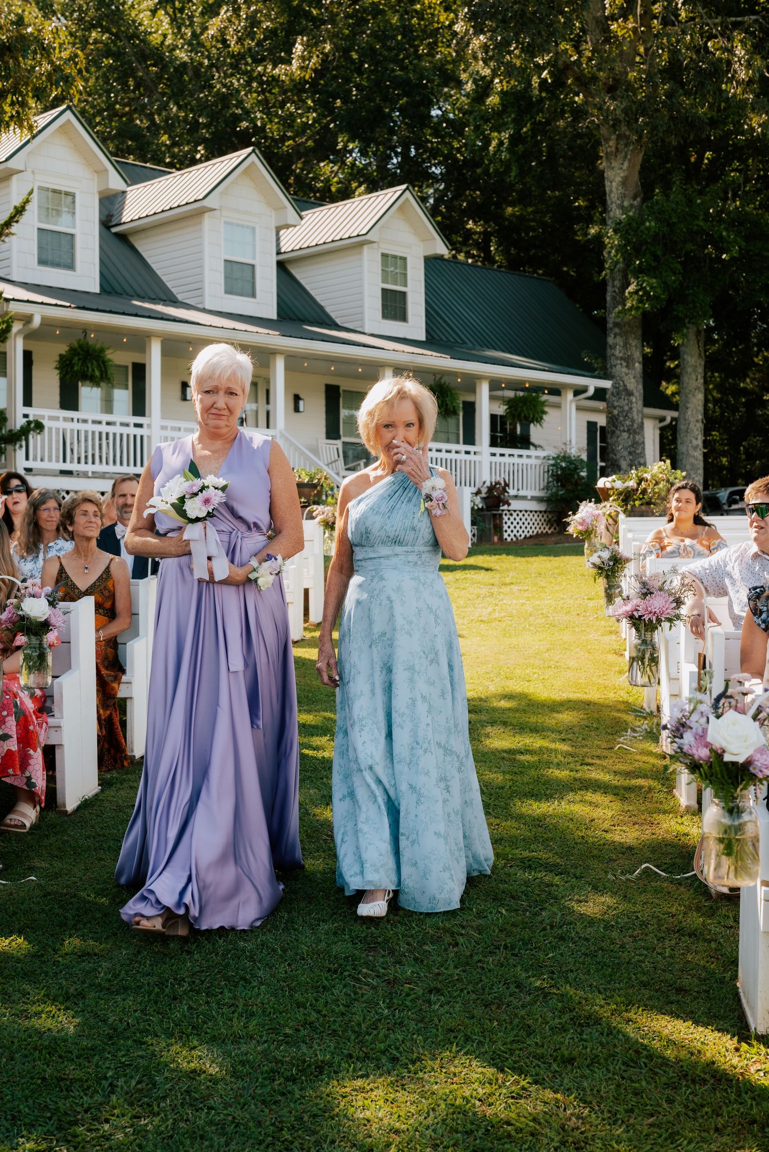 Outdoor wedding ceremony on a sunny lawn with two guests in formal purple and blue dresses walking down the aisle.