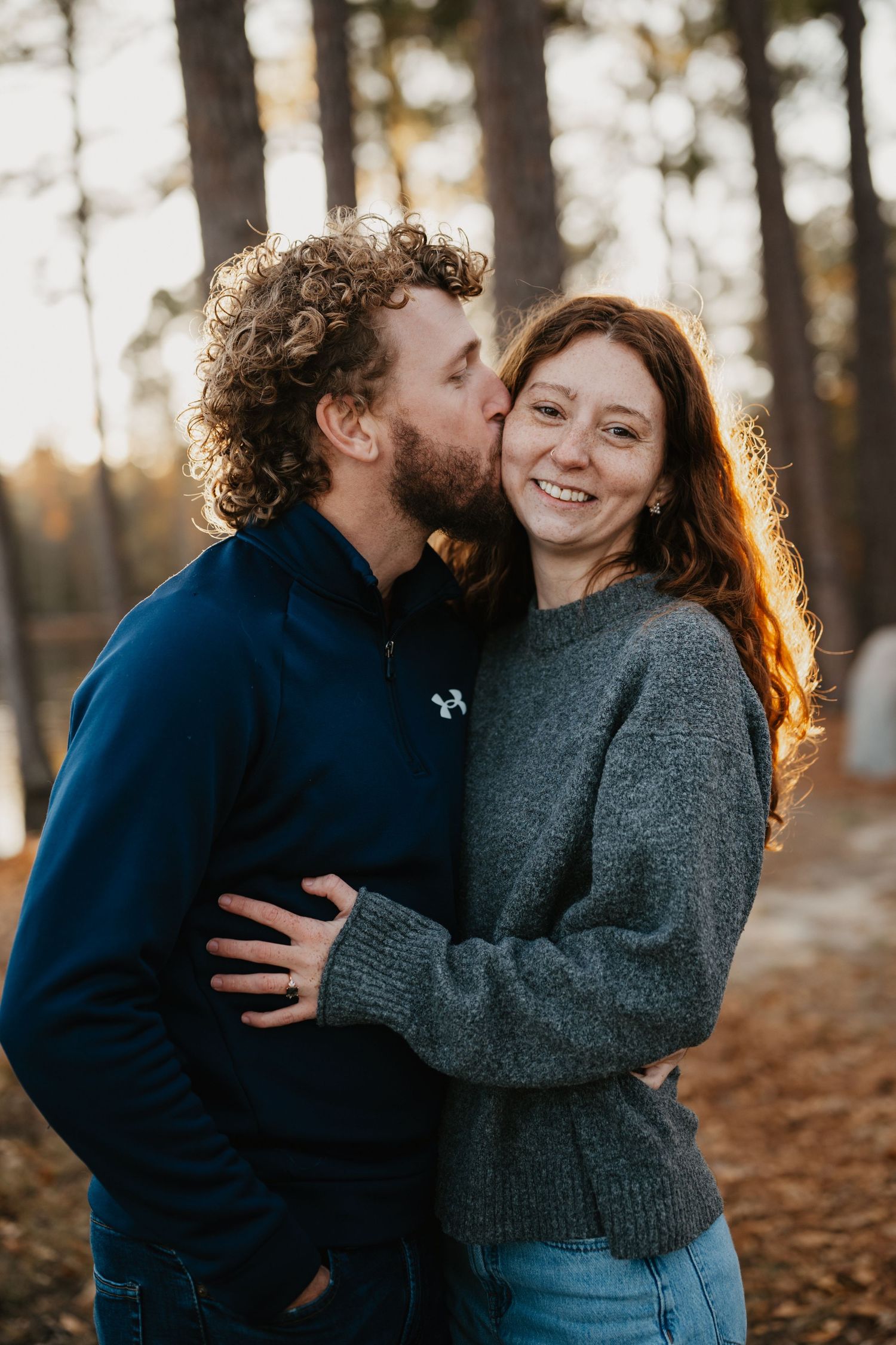 A couple in cozy sweaters share an intimate moment in a sunlit forest during golden hour.