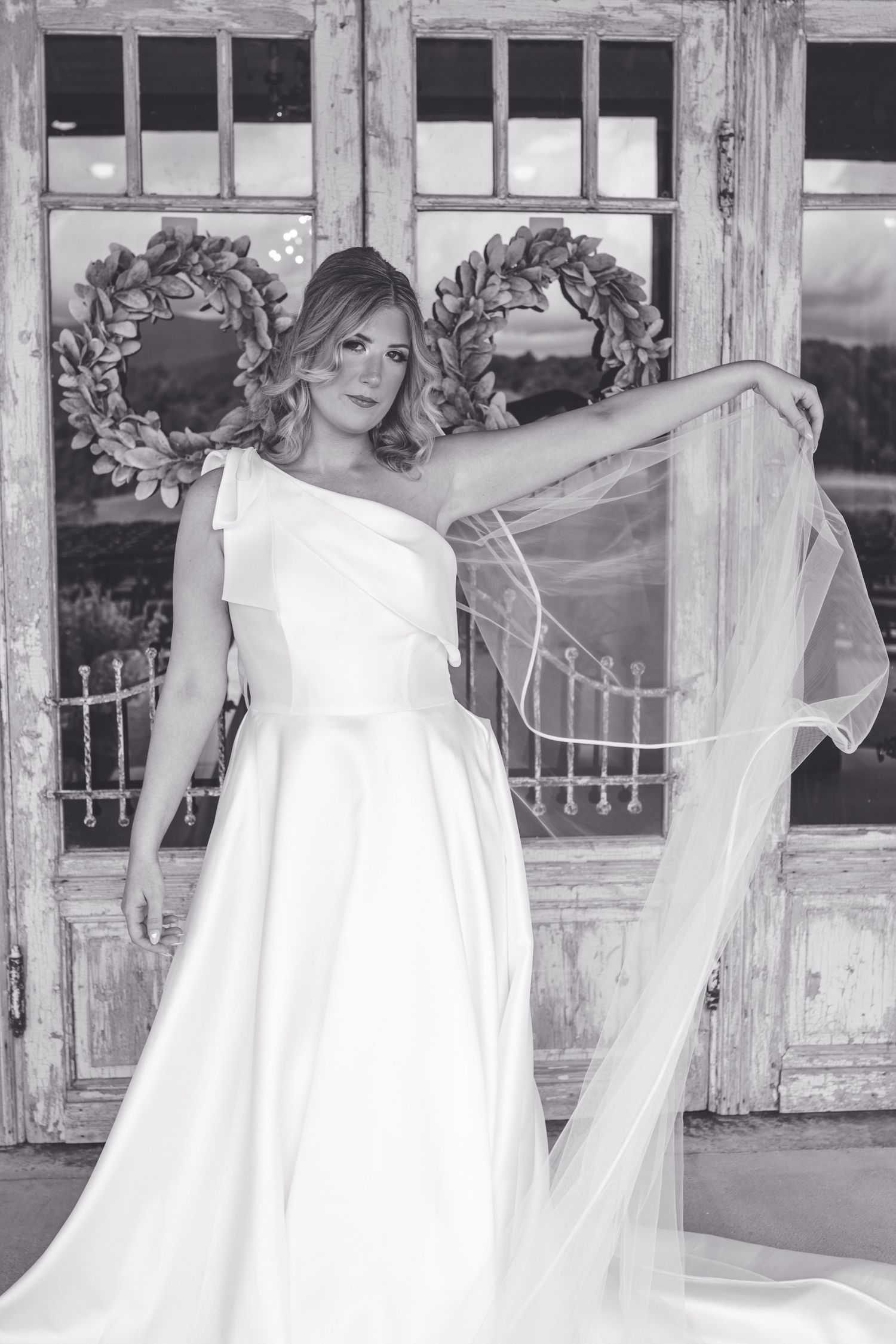 Bride in elegant one-shoulder white gown holds flowing veil in black and white wedding portrait.