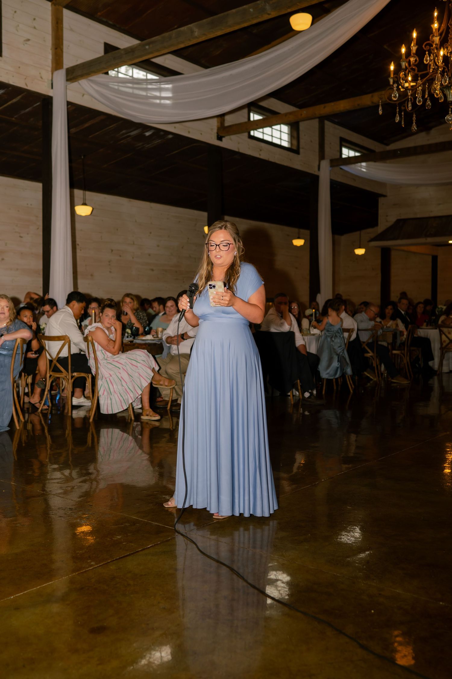 Bridesmaid in blue gown gives a speech at a rustic wedding reception with chandelier lighting.