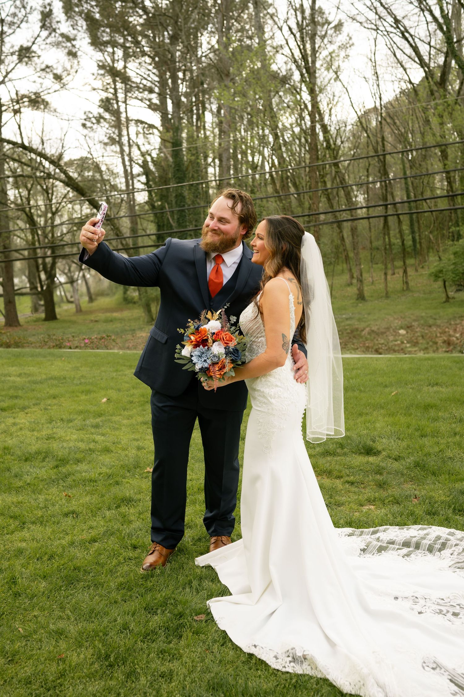 Newlywed couple taking a selfie outdoors, bride in white gown with veil holding colorful bouquet, groom in navy suit with red tie.