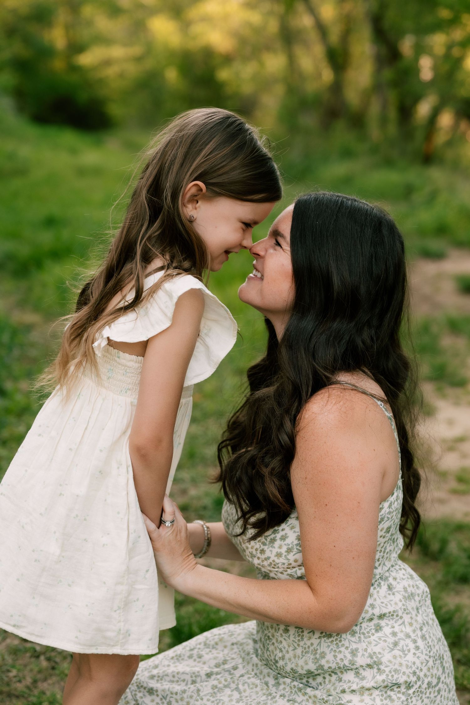 Mother and daughter share tender moments in nature wearing white dresses against a lush green backdrop.