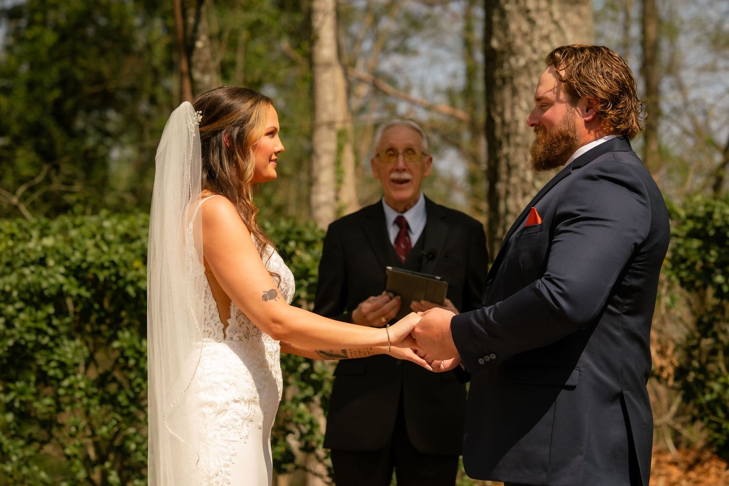 Bride and groom hold hands during outdoor wedding ceremony while officiant smiles behind them in a wooded setting.