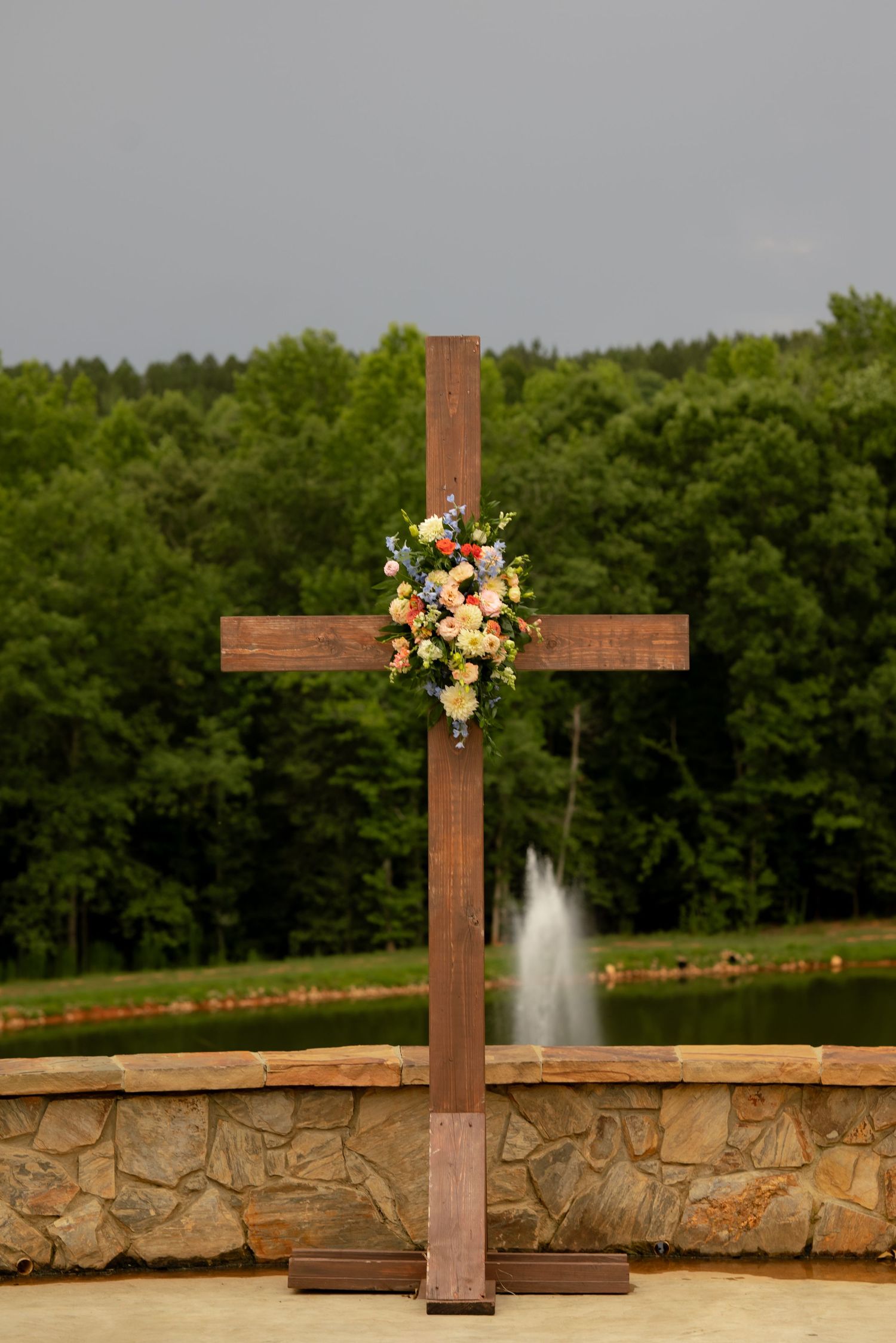 Wooden cross decorated with colorful floral arrangement at outdoor wedding ceremony, with pond fountain and green trees in background.