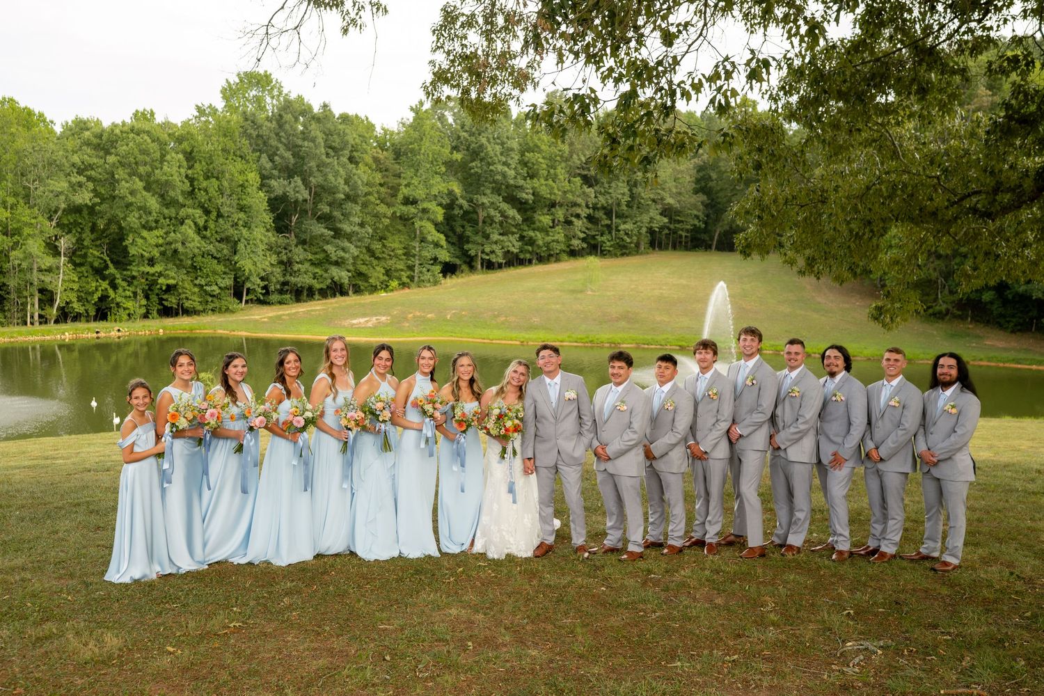 Wedding party in light blue dresses and gray suits posing on a green lawn with a pond and trees in background.