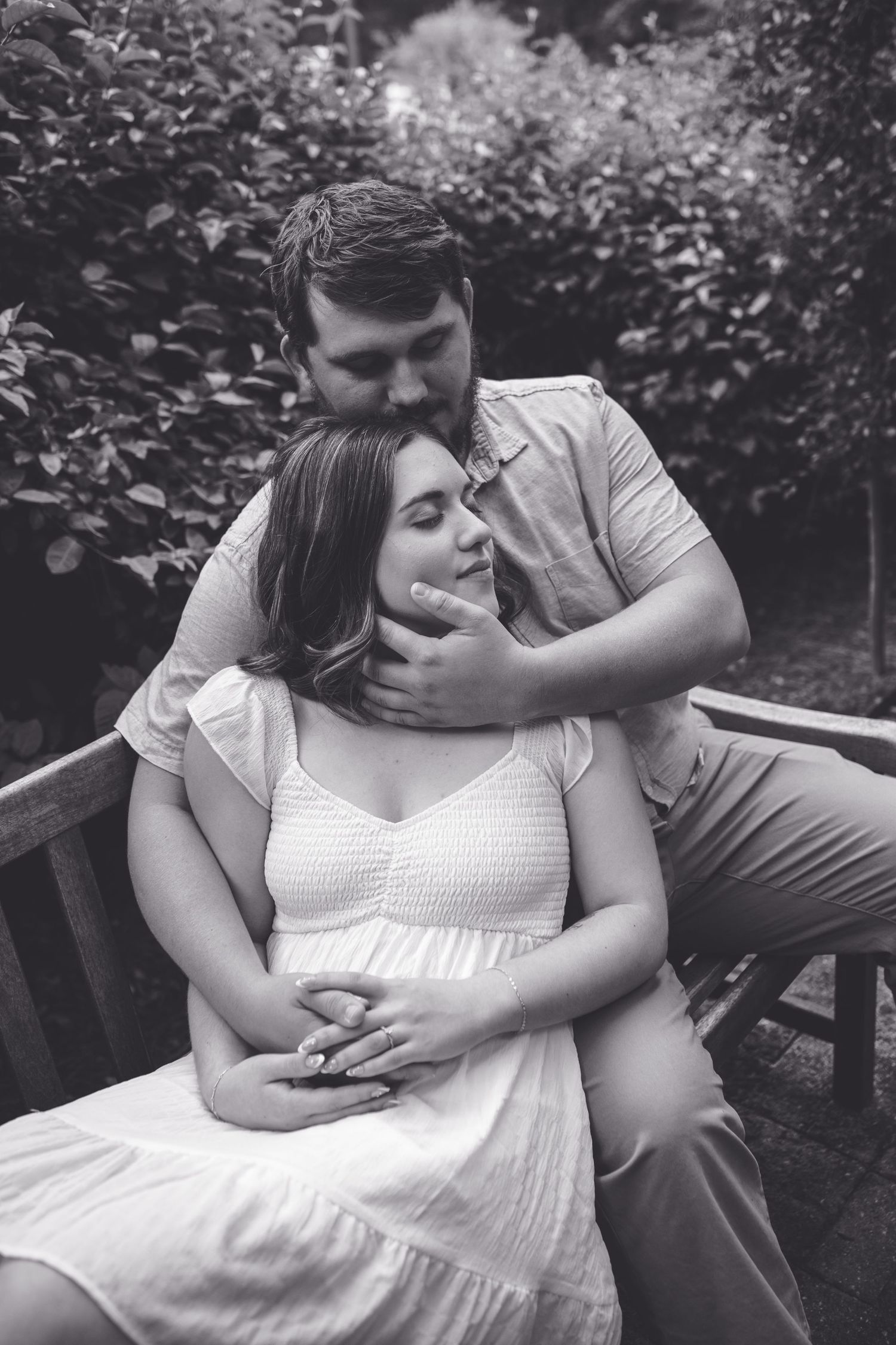 Black and white photo of a couple sharing a tender moment on a garden bench, woman in white dress cradling baby bump.