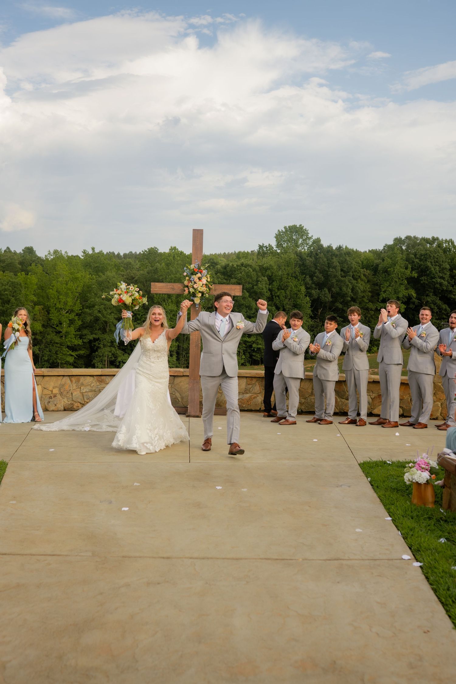 Bride and groom celebrate at outdoor wedding ceremony with wedding party beside rustic wooden cross.
