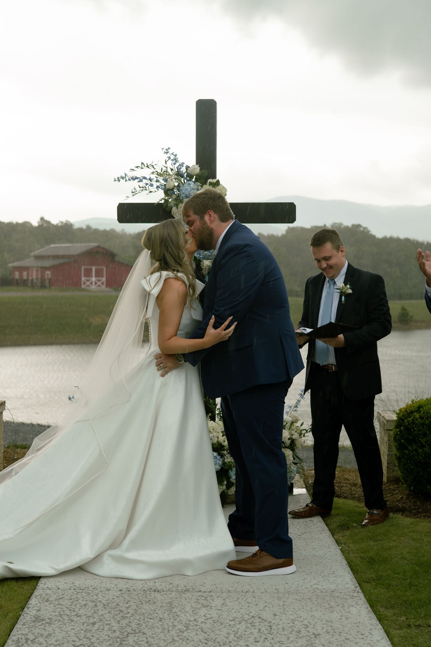 Bride and groom sharing first kiss at outdoor wedding ceremony in front of wooden cross with mountain backdrop.