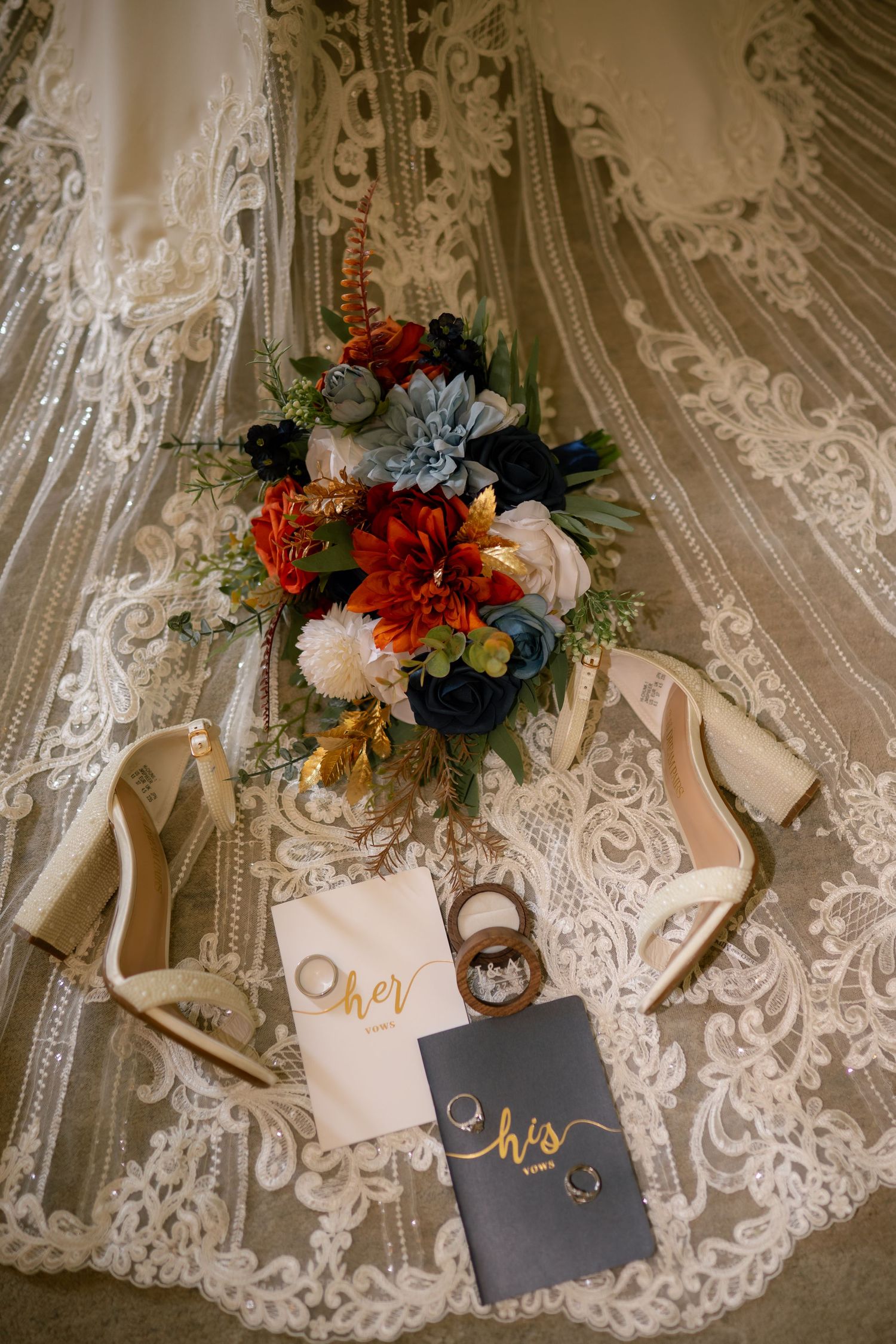Wedding flat lay featuring a lace bridal veil, colorful bouquet, gold heels, rings, and his and her vow books.