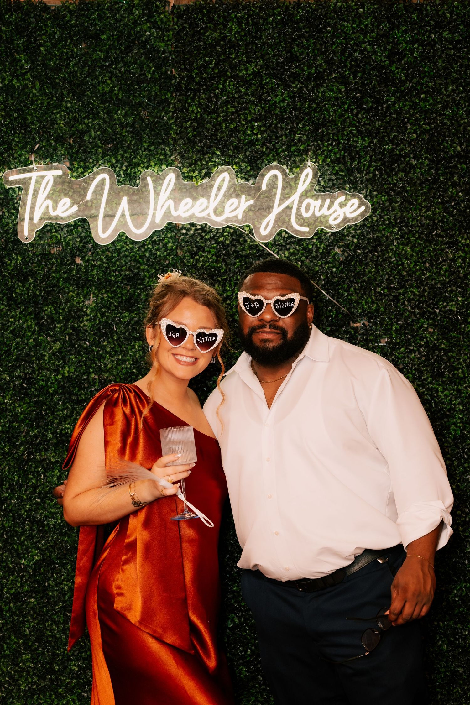 Couple in heart-shaped sunglasses pose before a green boxwood wall with The Wheeler House neon sign.