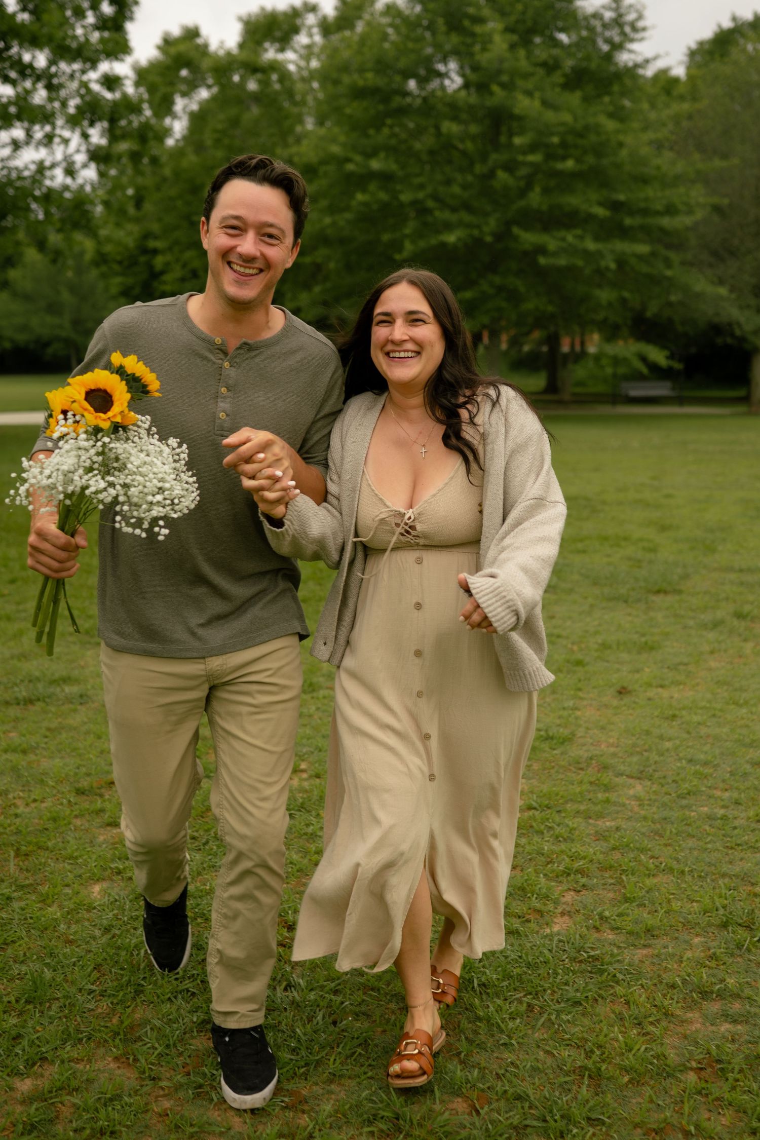 Happy couple walking in park, man holding sunflowers and baby's breath bouquet, woman in cream dress, smiling outdoors.