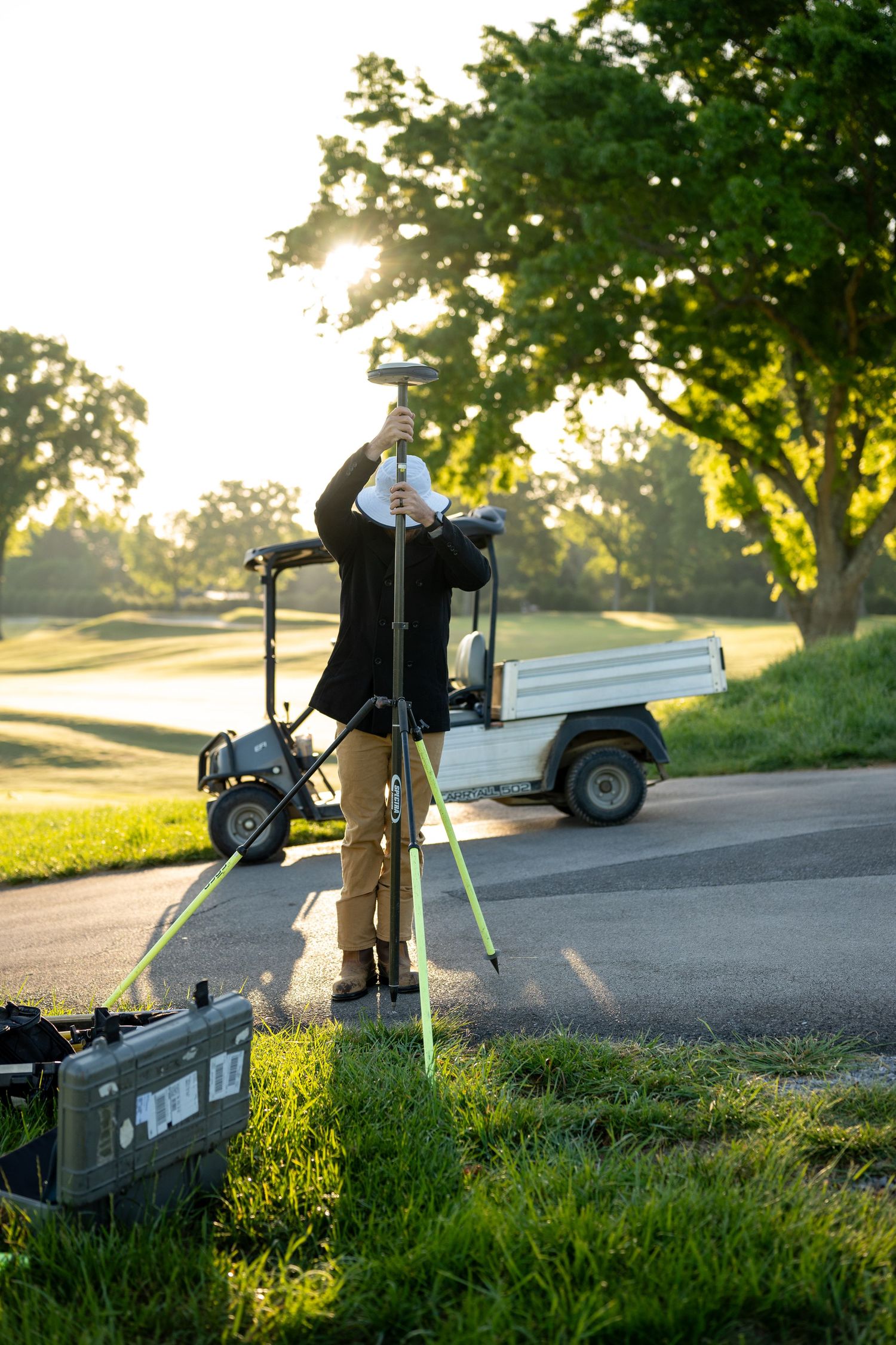 Surveyor using GPS equipment on a tripod at a sunny golf course, with a utility vehicle and lush green trees in the background.