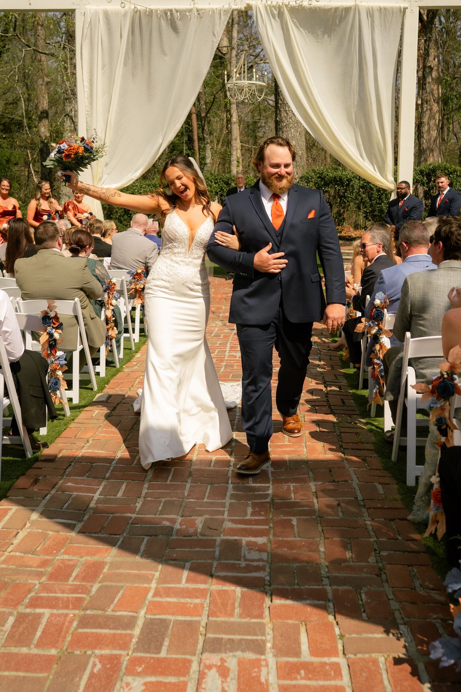 Joyful bride and groom walk down brick aisle after outdoor wedding ceremony under white draped arch.