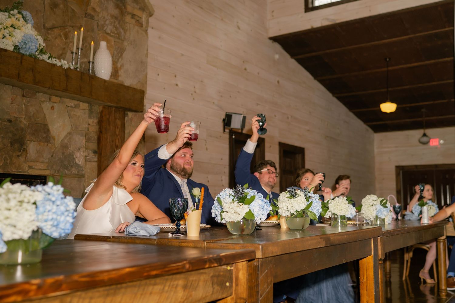 Wedding party raises glasses in a toast at the head table decorated with blue hydrangea centerpieces.