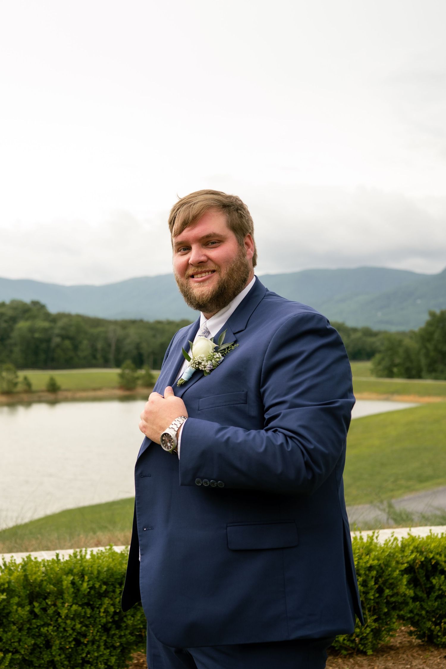 Smiling groom in navy blue suit with boutonniere poses outdoors with mountain and lake backdrop.