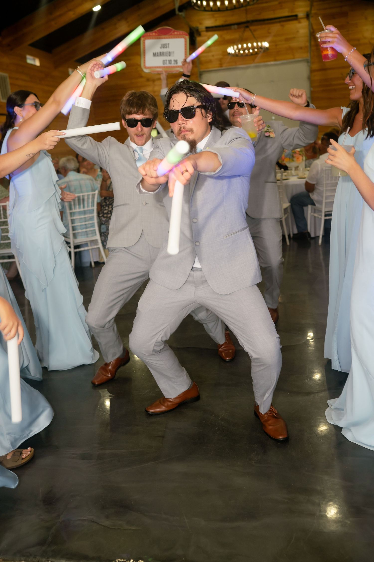 Groom dancing energetically with glow sticks at wedding reception surrounded by wedding party.