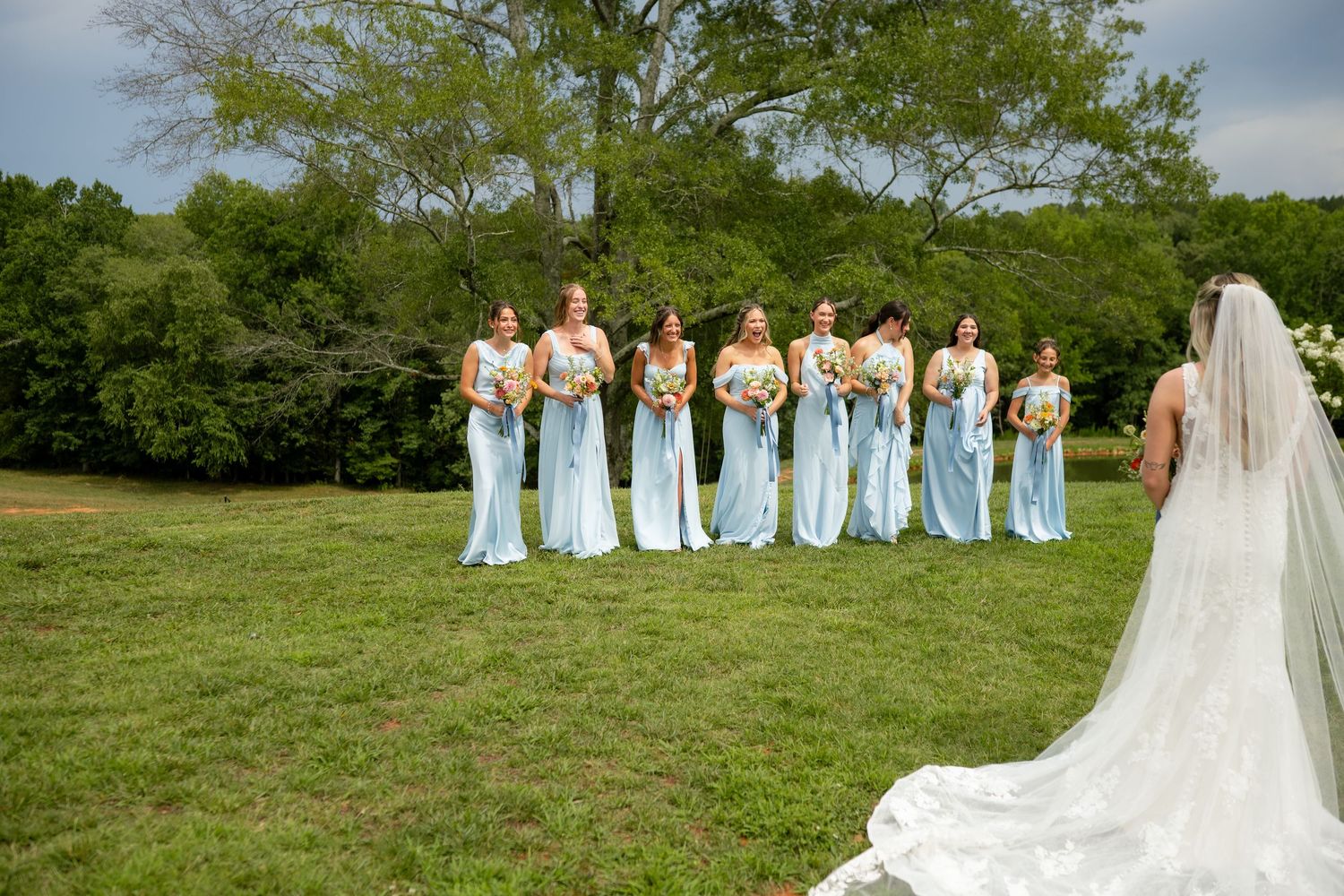 Bride in white gown faces bridesmaids in light blue dresses holding bouquets on green lawn.