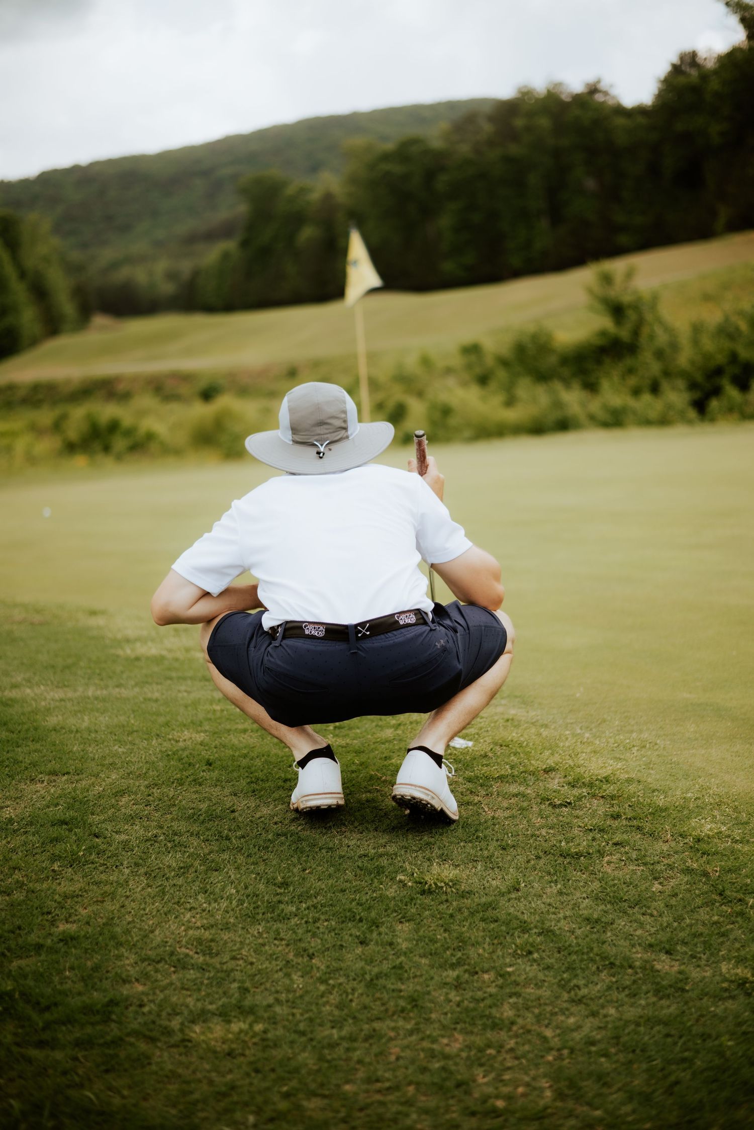 Golfer crouching on green in white shirt and hat, reading putt with flag in background on scenic course.