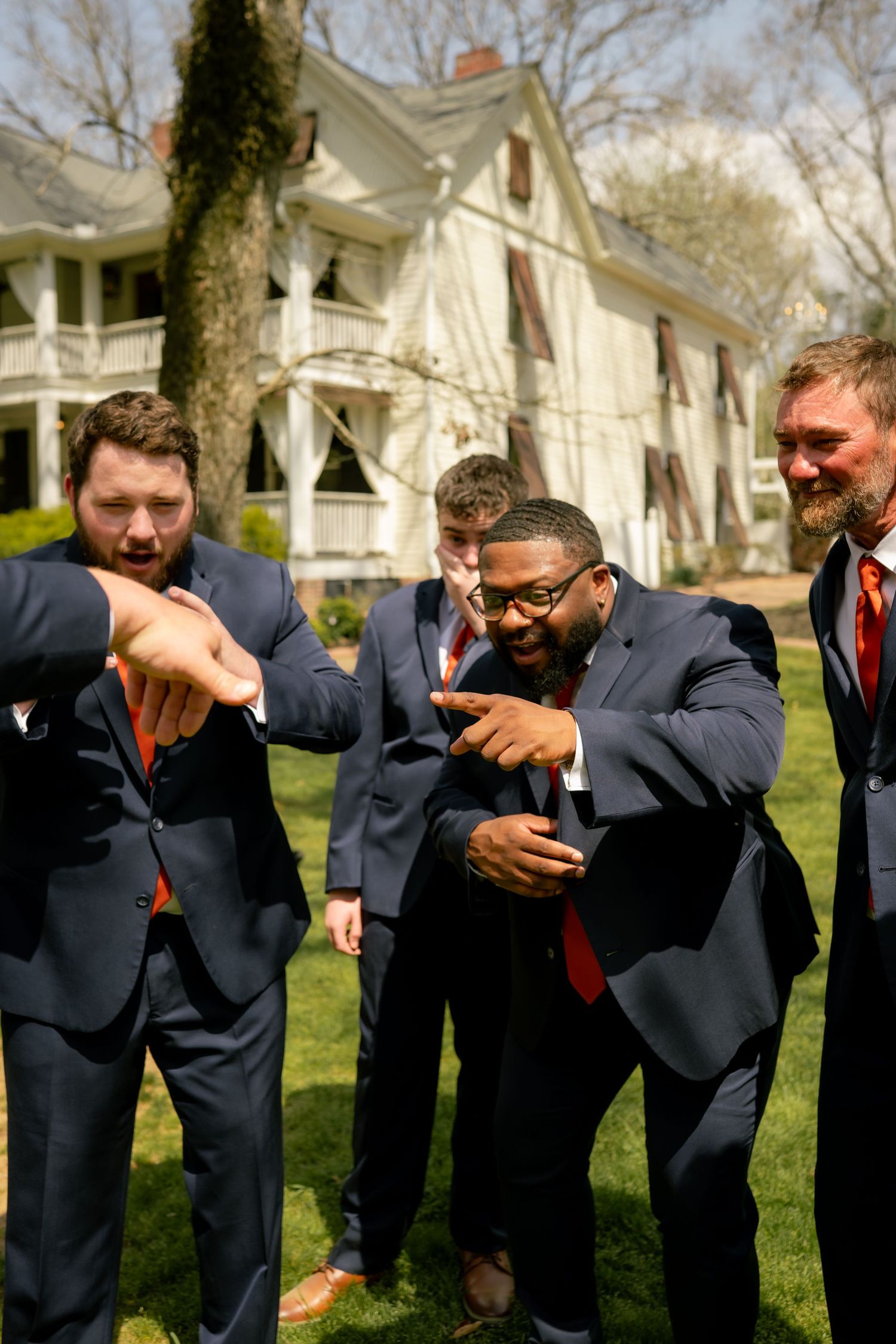 Groomsmen in navy suits with red ties laughing and pointing outdoors near a white historic house.