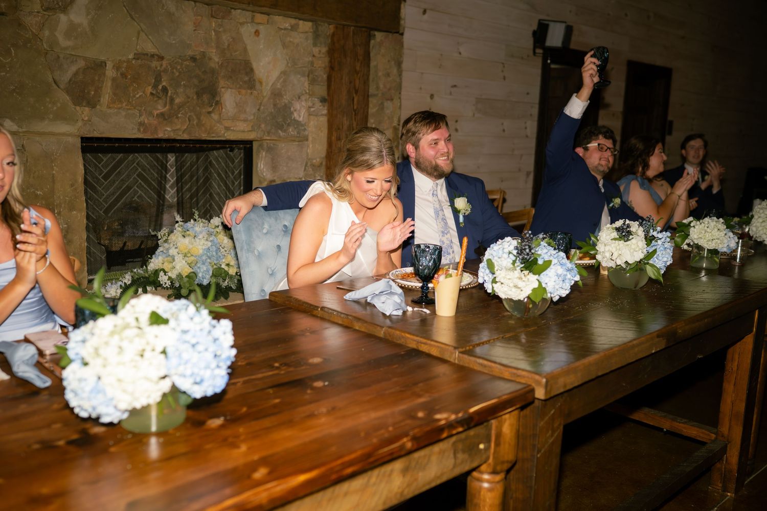 Bride and groom seated at rustic wooden head table during wedding reception.