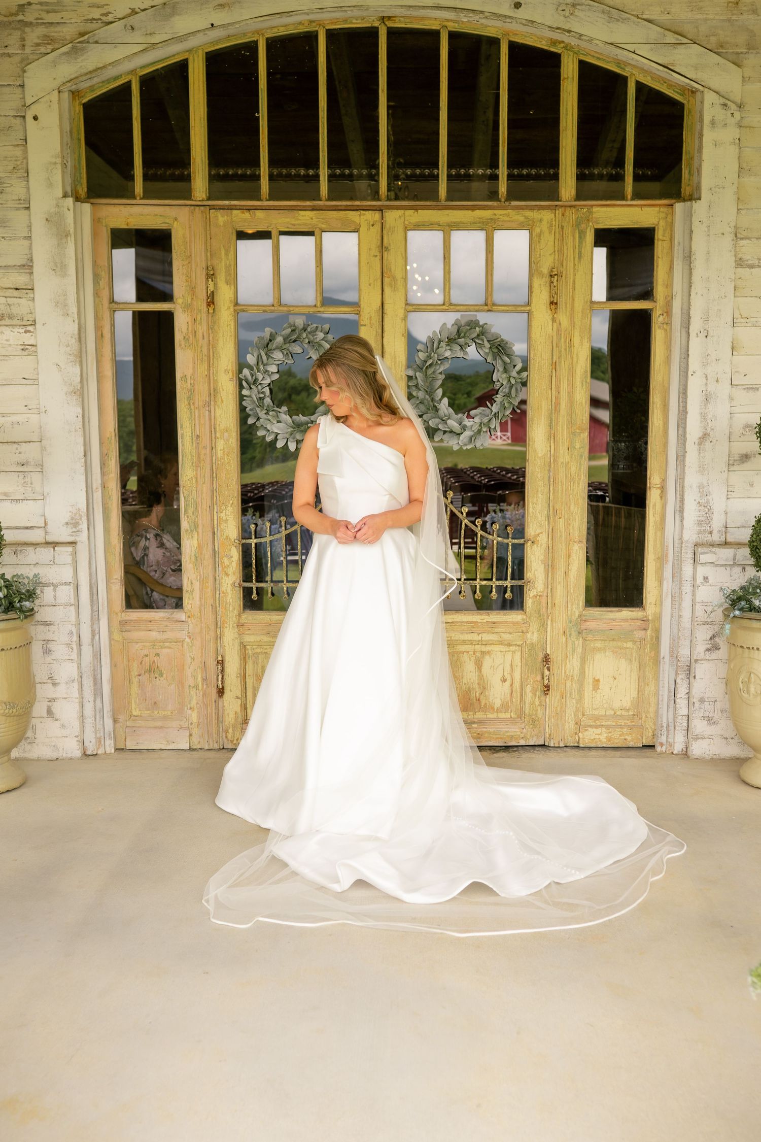 Bride in elegant one-shoulder white ball gown with long train stands before rustic French doors with wreath decor.