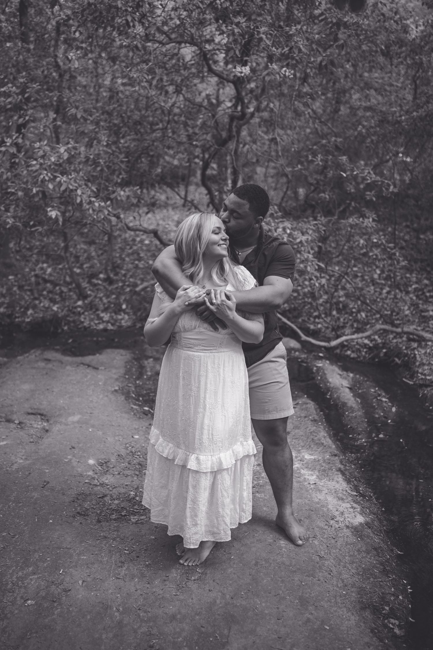 Black and white photo of a couple embracing outdoors, man hugging woman in white dress among trees.
