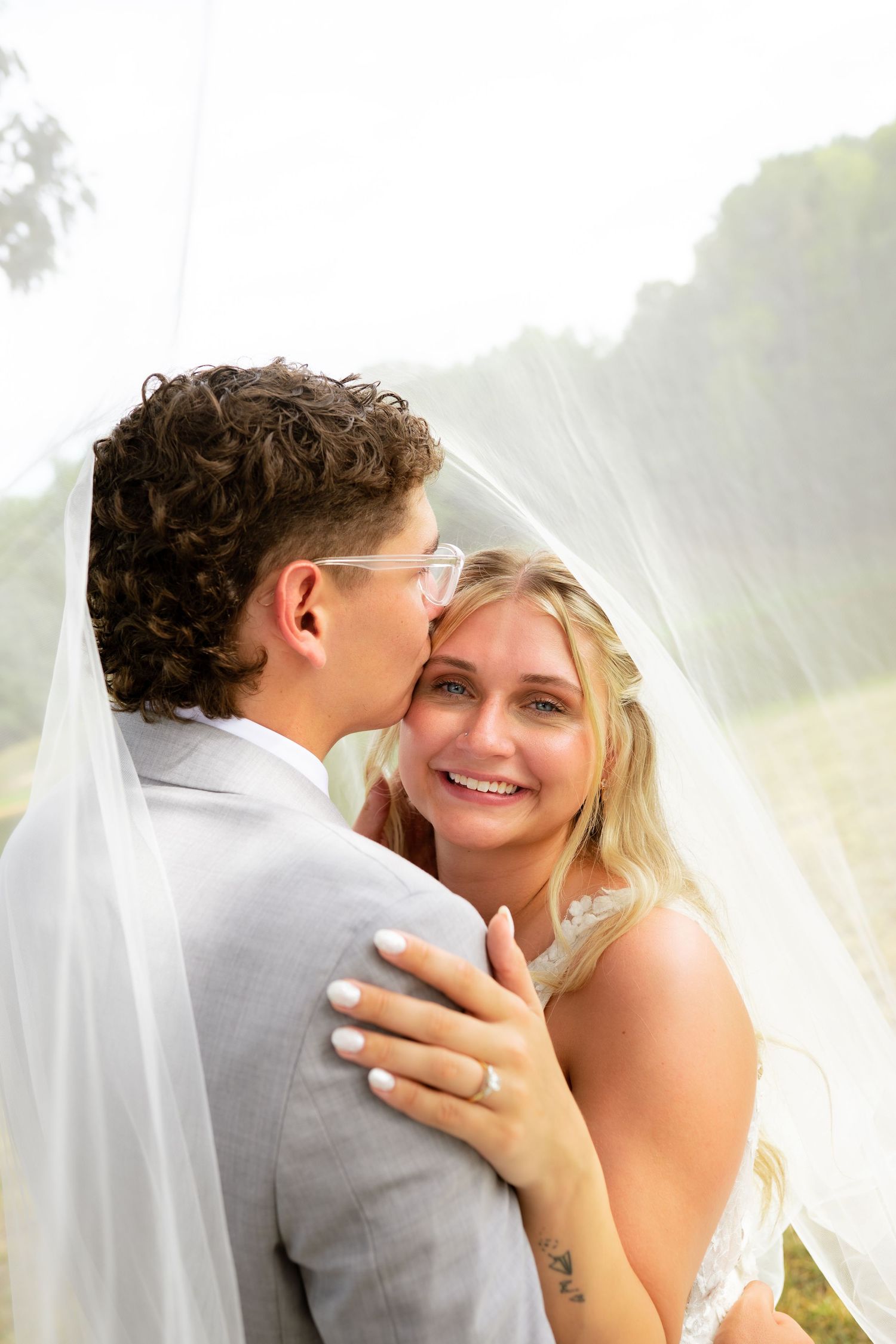 Groom kissing bride on the cheek under a flowing veil as she smiles during their wedding day.