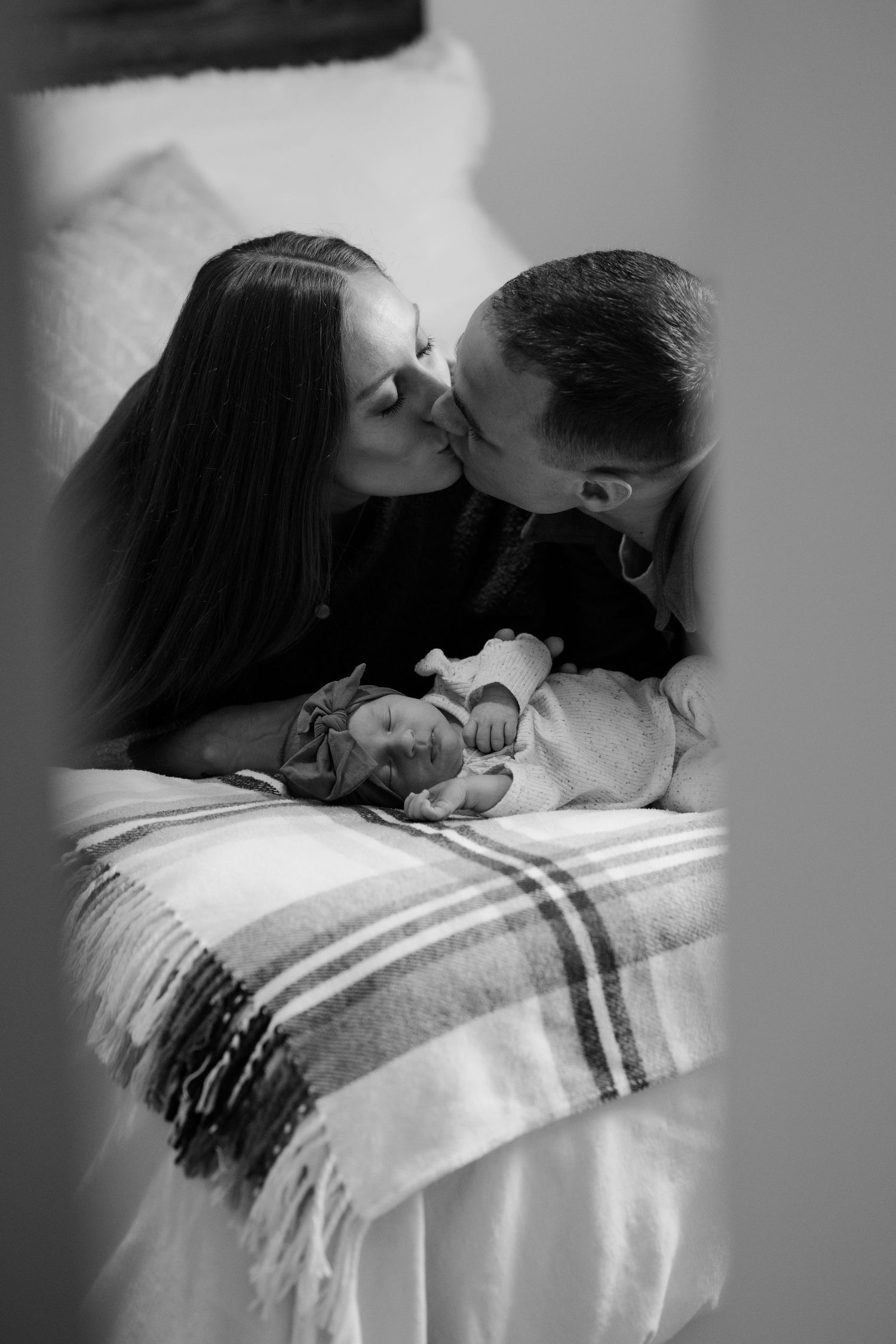 Intimate black and white portrait of parents kissing near sleeping newborn.