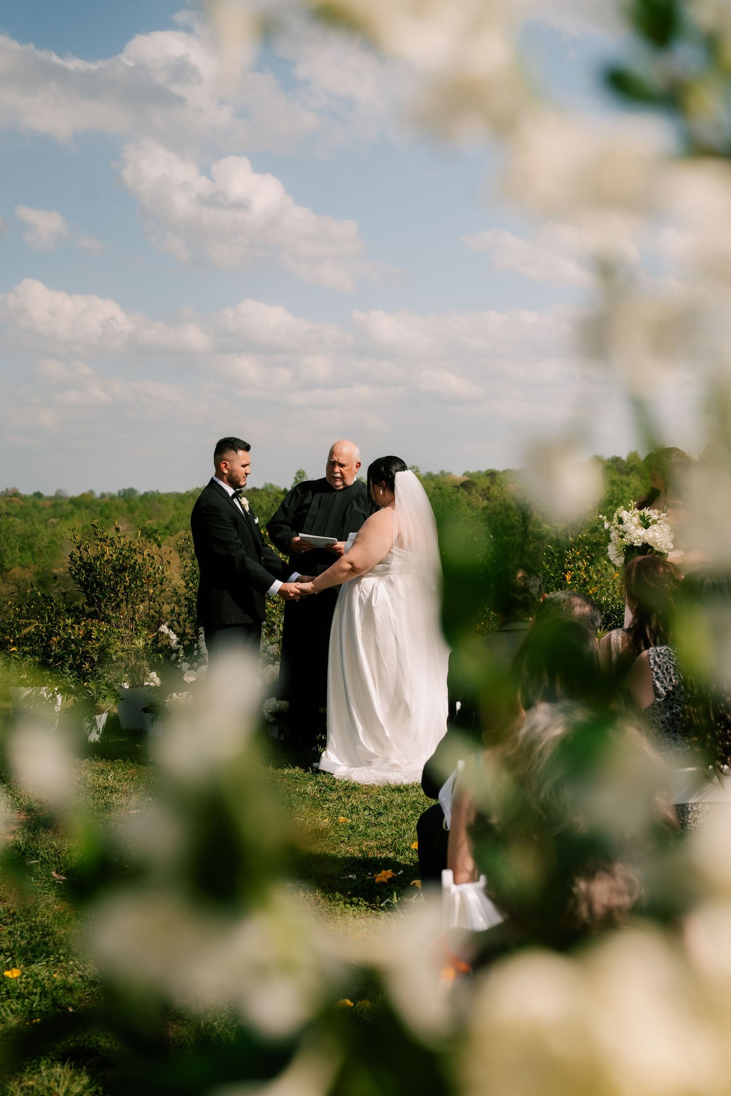 Outdoor wedding ceremony in a green field framed by delicate white blossoms and fluffy clouds in a blue sky.  North Carolina