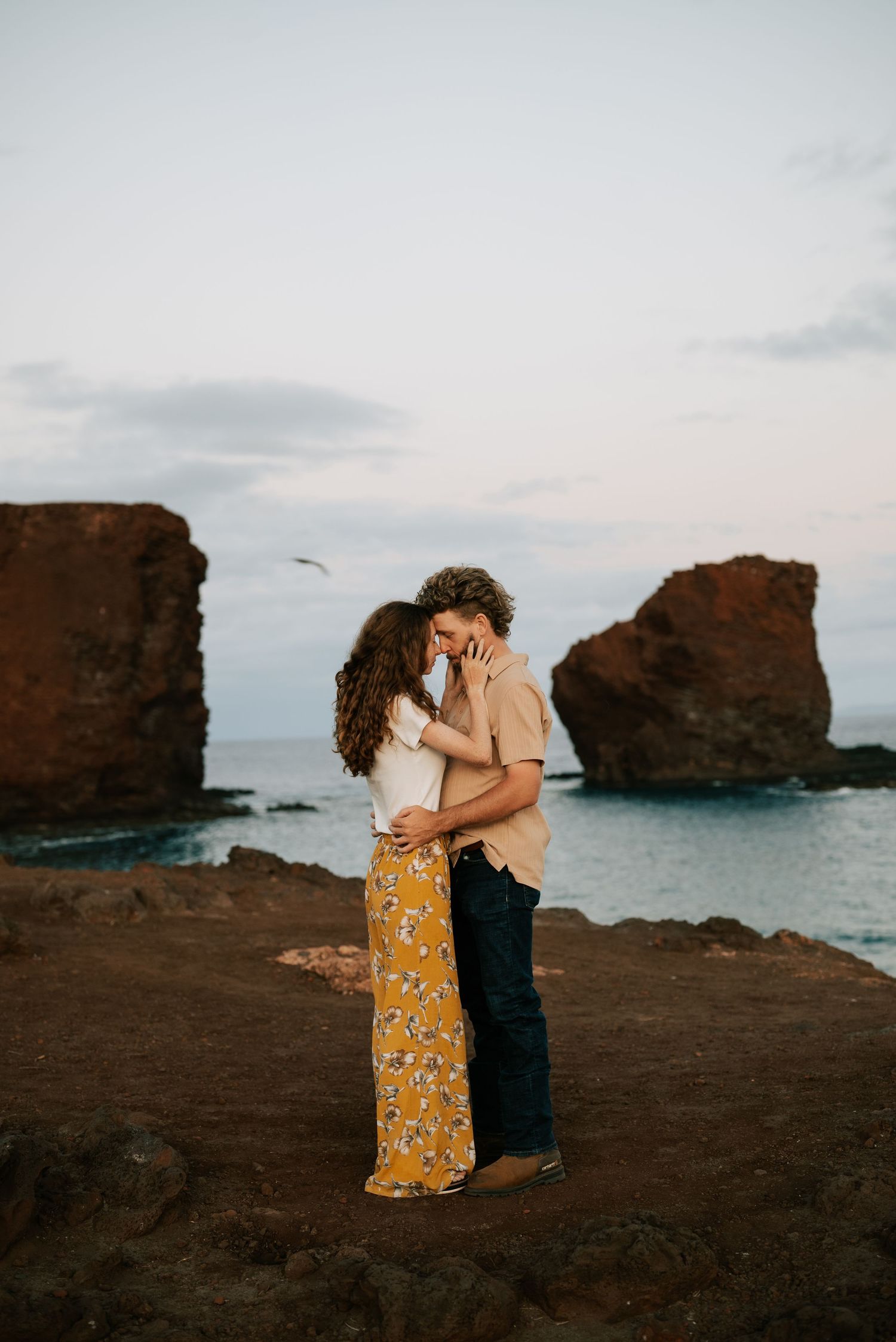 A couple embraces against a backdrop of rocky coastal cliffs and ocean during sunset.