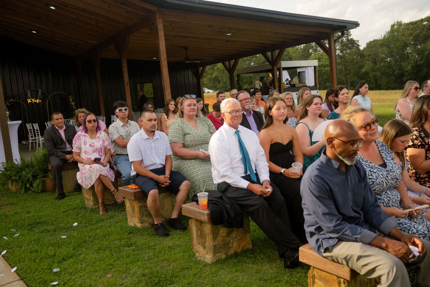 Guests seated on wooden benches at an outdoor wedding ceremony on a green lawn near a rustic barn venue.