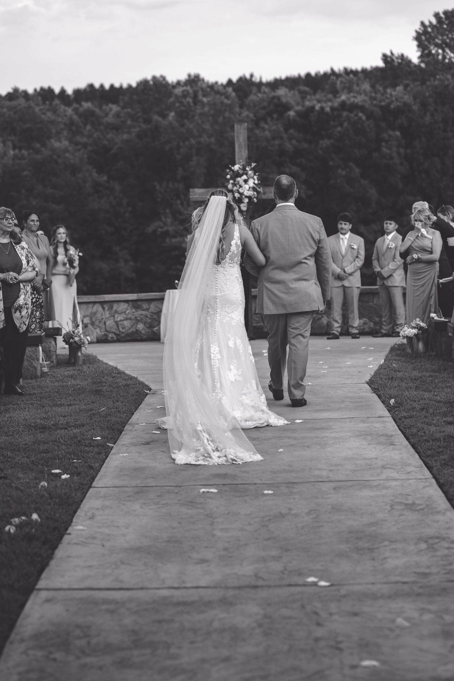 Black and white photo of a bride walking down the aisle with her father toward a cross at an outdoor wedding ceremony.