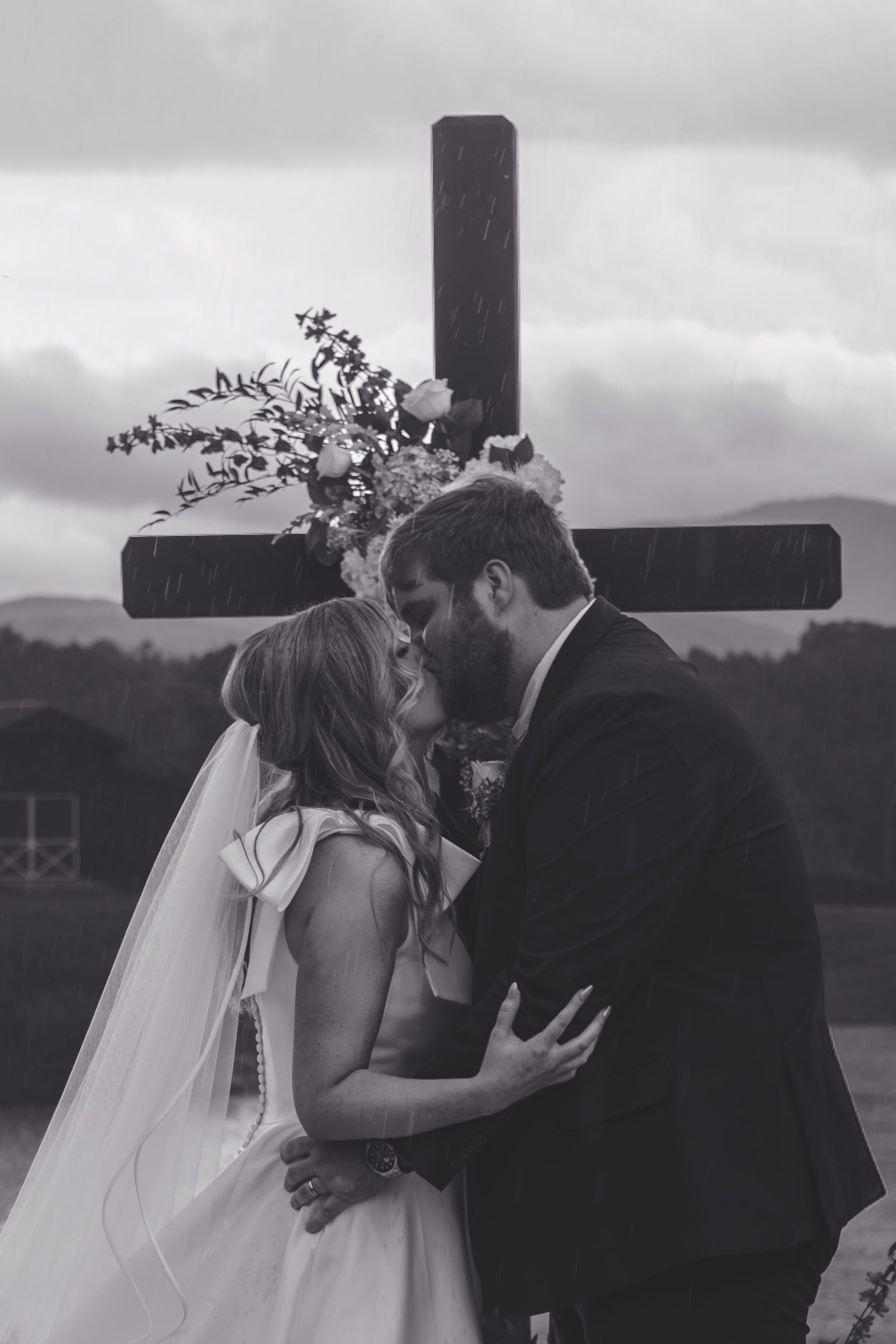 Black and white photo of a bride and groom sharing their first kiss in front of a floral-adorned wooden cross.