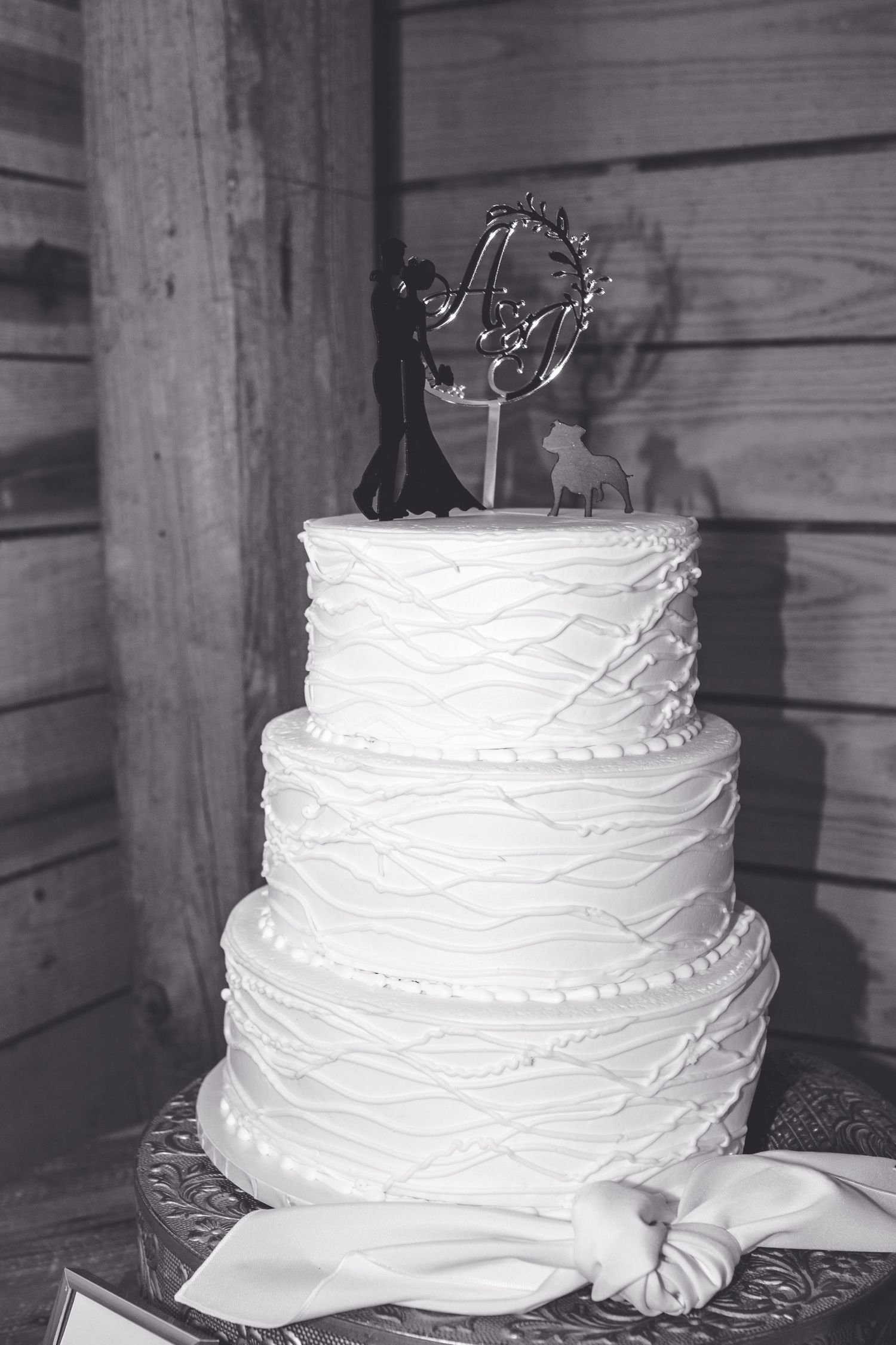 Three-tier white wedding cake with textured frosting and couple figurine topper on rustic wooden backdrop.