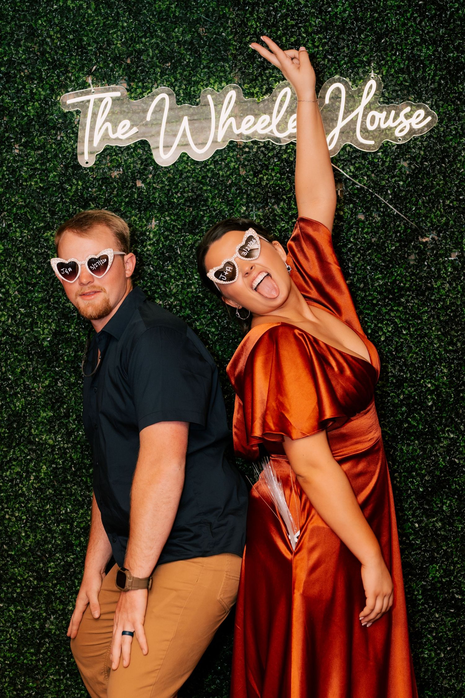 Couple in heart sunglasses pose playfully in front of The Wheeler House neon sign on green hedge wall.