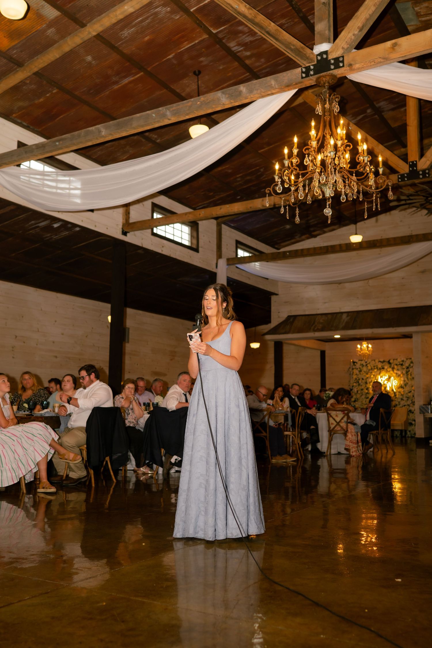 Bridesmaid in blue gown delivers a toast at an elegant barn wedding reception with wood ceiling.