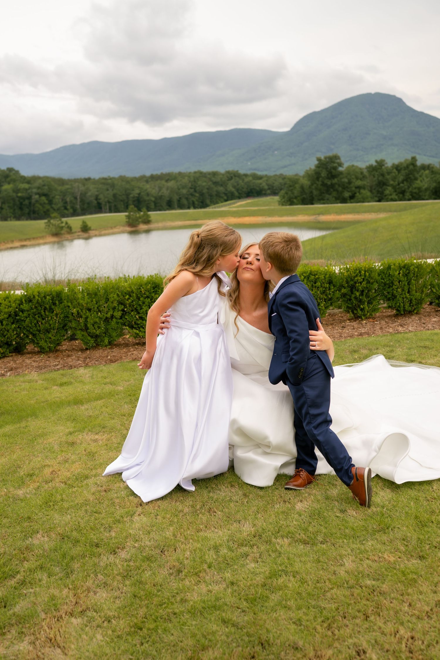 Bride in white gown kneels to kiss two children outdoors with scenic mountain and lake behind.