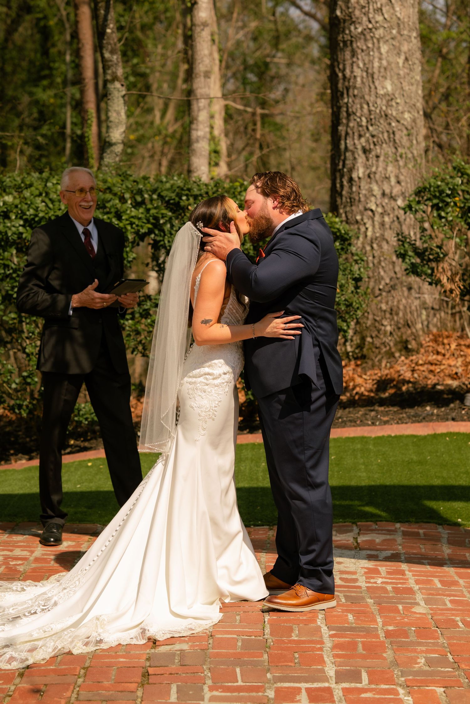 Bride and groom share first kiss at outdoor wedding ceremony on brick patio with officiant and lush greenery.